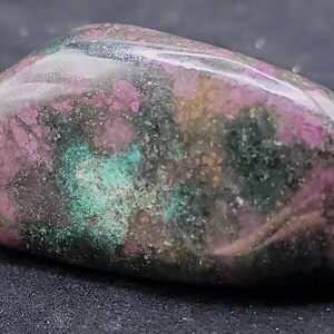 Puede incluir: Una piedra preciosa pulida con una mezcla de colores, incluyendo rosa, verde y marrón. La piedra tiene una forma suave y redondeada y una superficie ligeramente brillante. Los colores se distribuyen en un patrón moteado, creando una apariencia única y natural.