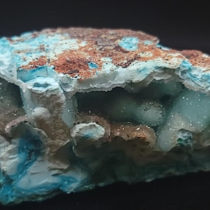May include: A close-up of a mineral specimen with a rough, textured surface. The mineral features a mix of colors, including light blue, brown, and hints of green. The overall shape is irregular, with visible crystalline structures.