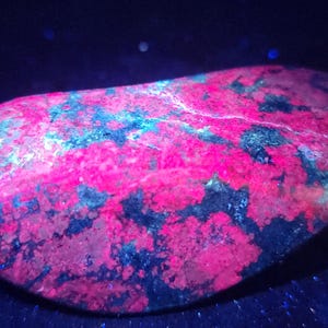 May include: A polished, irregularly shaped stone with a vibrant colour palette. The surface displays a striking combination of hot pink, deep blue, and black hues, creating a visually dynamic and textured appearance. The stone is set against a dark background.