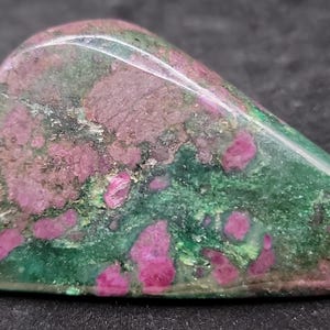 Puede incluir: Primer plano de una piedra preciosa pulida. La piedra presenta una base de color verde oscuro con parches de rosa vibrante y un toque de verde más claro. La superficie es lisa y reflectante, mostrando los patrones naturales.
