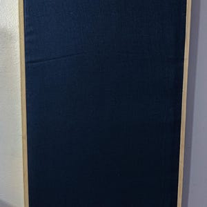 May include: A rectangular, navy blue fabric panel is set within a light wood frame. The fabric has a slightly textured appearance. The frame is a light, natural wood color. The panel is mounted on a white wall.