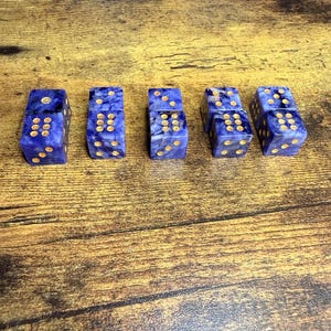 May include: Five blue dice with gold-colored dots are arranged on a wooden surface. The dice have a marbled appearance, with a mix of light and dark blue hues. The dots are evenly spaced and clearly visible, creating a classic game piece.
