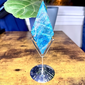 May include: A vibrant blue crystal point, mounted on a gold-colored metal stand with a circular base. The crystal has a textured surface and is displayed on a wooden table. The background includes a green plant and a blue pot.