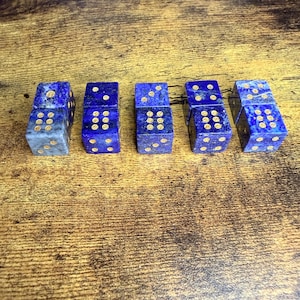 May include: Six blue dice with gold-colored dots are arranged on a wooden surface. The dice are made of a dark blue stone with gold-colored dots. The dice are used for games and are a unique gift idea.