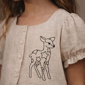 May include: A light beige linen button-down dress with short, puffed sleeves. The dress features a black embroidered deer with star-shaped markings. The dress has a square neckline and is buttoned down the front.