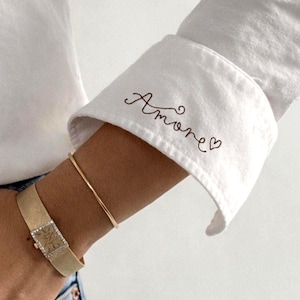 May include: White button-down shirt with the word "Amore" embroidered on the cuff. A gold watch and a thin gold bracelet are visible on the wrist. The person is wearing blue jeans.