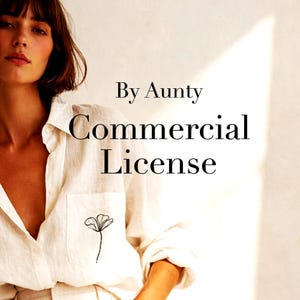 May include: A white button-down shirt featuring a small black line-art flower illustration on the chest pocket. The text By Aunty Commercial License is printed in black over the shirt. The garment has a collar and long sleeves with rolled cuffs.