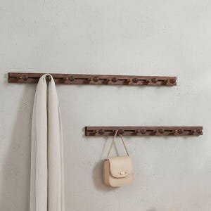 Puede incluir: Dos percheros de madera con múltiples pomos redondos. El perchero superior sostiene una bufanda de color claro, y el perchero inferior sostiene un pequeño bolso beige. Los percheros están montados en una pared gris claro.