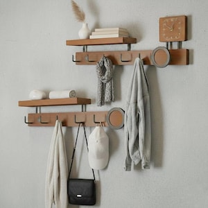 May include: Wooden wall-mounted coat rack with shelves, hooks, and a clock. The rack is made of light brown wood with metal accents. A gray scarf, jacket, hat, and purse hang from the hooks. Decorative items are on the shelves.