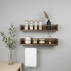 May include: Two dark wood floating shelves with brass accents are mounted on a gray wall. The top shelf holds glass jars with wooden lids and a black vase. The bottom shelf has a towel rack with a white towel and decorative jars.