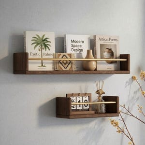 Könnte beinhalten: Zwei schwebende Regale aus dunklem Holz mit goldenen Metallschienen. Das obere Regal enthält Bücher mit den Titeln Exotic Palms, Modern Space Design und Artisan Forms sowie eine beige Vase. Das untere Regal trägt einen Klappkalender.