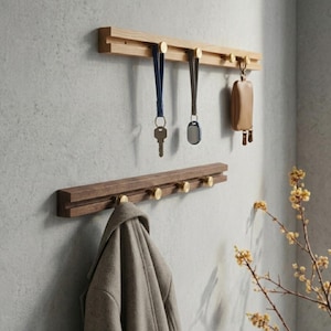 Puede incluir: Dos percheros de pared de madera con ganchos de latón. El perchero superior es de madera clara, con llaves y un llavero. El perchero inferior es de madera oscura, con un abrigo. Una ramita de flores amarillas está en el fondo.