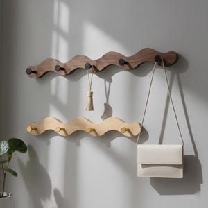 May include: Two wooden coat racks with a wave-like design, mounted on a wall. The top rack is dark brown with black knobs, the bottom is light wood with gold knobs. A white handbag and a tassel hang from the racks.