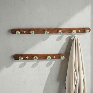 Puede incluir: Dos percheros de pared horizontales hechos de madera marrón oscura. Cada soporte cuenta con varios ganchos circulares de tono latón con incrustaciones de piedra veteada verde. Una prenda beige cuelga de uno de los ganchos superiores.