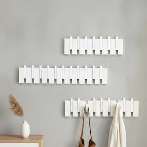 Op de afbeelding: Drie witte houten wandkapstokken met een minimalistisch ontwerp. Elke kapstok heeft een reeks rechthoekige pinnen. Eén kapstok houdt een bruine handtas en een witte jas vast. Ze zijn gemonteerd op een lichtgrijze muur.