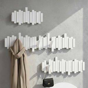 May include: Three white wooden coat racks mounted on a gray wall. The racks are made of vertical rectangular pieces of wood. A tan coat hangs on one rack, and a black purse hangs on another.