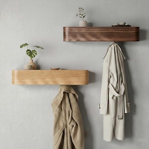 Puede incluir: Dos estantes flotantes de madera en tonos marrón claro y oscuro están montados en una pared gris. Cada estante tiene un frente curvo y sostiene pequeños objetos decorativos. Debajo de los estantes cuelgan abrigos, agregando un elemento funcional y elegante.