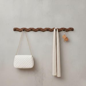 Puede incluir: Un perchero de pared de madera marrón oscuro con un diseño ondulado. Un bolso blanco acolchado con una cadena dorada, una bufanda color crema y unas llaves cuelgan del perchero. El perchero está montado en una pared gris claro.
