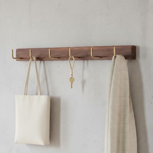 May include: A wooden wall-mounted coat rack with six brass hooks. A cream tote bag, a key with a leather fob, and a beige scarf hang from the hooks. The rack is made of dark wood and is mounted on a light grey wall.
