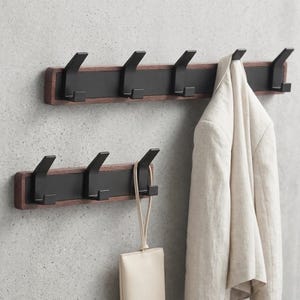 May include: Two wooden coat racks with black metal hooks mounted on a gray wall. One rack has a light-colored jacket hanging from it, and the other has a small, light-colored purse hanging from it. The wood is a dark brown color.