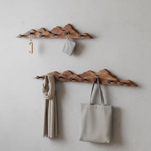 May include: Two wooden mountain-shaped coat racks with dark metal hooks. One rack holds a key and a small pouch, the other a scarf and a tote bag. The racks are mounted on a light gray wall.