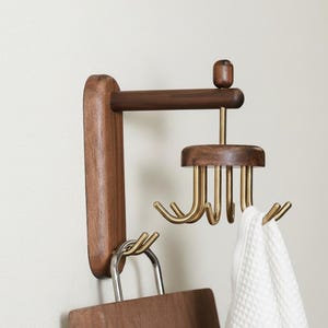 May include: A wooden wall-mounted organiser with multiple hooks. The organiser features a dark brown wooden base and a horizontal arm with brass hooks. A white towel hangs from one of the hooks. The design is minimalist and modern.