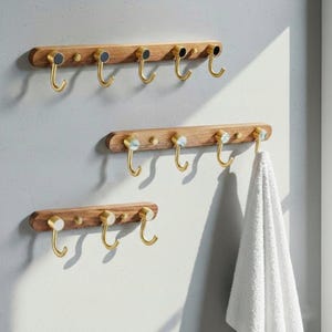 Könnte beinhalten: Drei Garderoben aus Holz mit Messinghaken sind an einer hellen Wand befestigt. Die obere Leiste hat sechs Haken mit schwarzen Akzenten, die mittlere Leiste hat fünf Haken mit weißen Akzenten und die untere Leiste hat drei Haken mit weißen Akzenten. Ein weißes Handtuch hängt an einem der Haken.