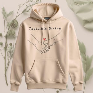 Könnte beinhalten: Beige Kapuzenpullover mit Fronttasche und Kordelzugkapuze. Der Kapuzenpullover zeigt den Text "Invisible String" über einer Strichzeichnung von zwei Händen, die sich halten, mit einem kleinen roten Herz dazwischen. Der Hoodie ist aus weichem Material.