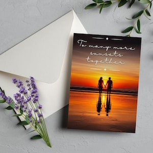 May include: Greeting card with a sunset beach scene, featuring two silhouetted figures holding hands. The card reads "To many more sunsets together" in elegant script. An open white envelope and lavender sprig are next to the card.