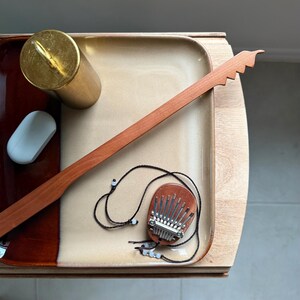 May include: A small wooden kalimba pendant with metal tines and a black cord, a long thin wooden stick with a serrated end, a gold-colored cylindrical container, and a small white oval object arranged on a rectangular two-toned ceramic tray.