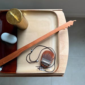 May include: A small wooden kalimba pendant with metal tines on a black cord, a long wooden stick with a serrated end, a gold-colored cylindrical weight, and a smooth white oval object, all arranged on a rectangular two-toned ceramic tray.