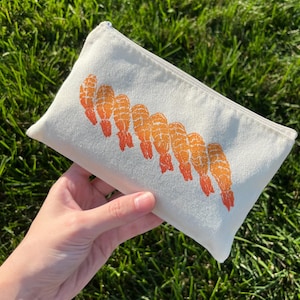 May include: A rectangular off-white canvas zippered pouch featuring a repeating graphic print of orange and red shrimp arranged in a diagonal line across the center. The fabric has a visible woven texture and a silver metal zipper closure.