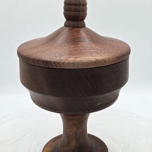 May include: A handcrafted wooden container with a lid. The container is made of dark brown wood and has a pedestal base. The lid has a rounded top with a finial. The container is likely used for storage or display.