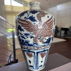 Puede incluir: Un jarrón de cerámica con base blanca, adornado con un diseño de fénix en tonos marrón y azul. El jarrón presenta motivos florales y un diseño detallado en la base. Tiene un cuello estrecho y un cuerpo bulboso.