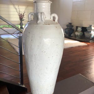 May include: A tall, off-white ceramic vase with a narrow neck, two handles, and a lid. The vase has a textured surface with brown speckles and a brown base. The vase is likely for decorative purposes.