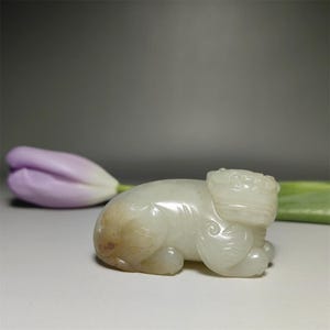 Puede incluir: Una figura de jade tallada en color marfil de una bestia mítica reclinada. La escultura tiene detalles y una superficie lisa y pulida. La figura se encuentra sobre un fondo suave y neutro, con un tulipán morado en el fondo.