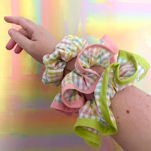 May include: A fabric hair scrunchie featuring a multi-colored pastel gingham pattern in shades of yellow, green, blue, and pink. The scrunchie is accented with ruffled edges trimmed in solid light pink and bright lime green fabric piping.
