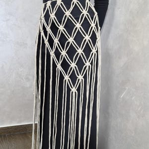 以下が含まれることがあります： クリーム色のマクラメのオーバーレイが付いた黒いスカート。マクラメはウエストにダイヤモンド模様があり、長いフリンジのディテールが特徴です。スカートは無地で、柔らかい生地で作られているようです。