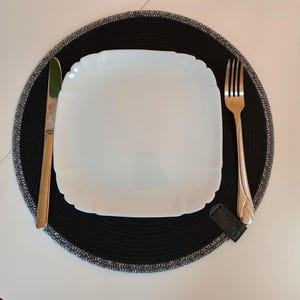 Könnte beinhalten: Ein Gedeck mit einem weißen quadratischen Teller, einer goldenen Gabel und einem Messer sowie einem schwarzen Platzset mit grauem Rand. Weitere schwarze Platzsets sind im Hintergrund zu sehen. Das Gedeck befindet sich auf einer weißen Oberfläche.