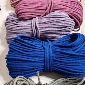 May include: A collection of colorful macrame rope in shades of purple, pink, blue, and gray. The rope is tightly wound into bundles, showcasing the texture and color variations. Ideal for crafting and decorative projects.