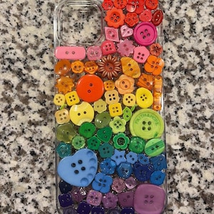 May include: A clear phone case decorated with a variety of colorful buttons. The buttons are arranged in a rainbow pattern, with red, orange, yellow, green, blue, and purple hues. Some buttons have the words "Faith & Hope".