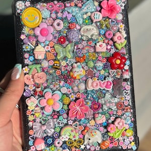 Könnte beinhalten: Eine schwarze Tablet-Hülle, die mit farbenfrohen Verzierungen bedeckt ist. Die Hülle ist mit verschiedenen Charms verziert, darunter Blumen, Schleifen, ein Smiley und das Wort "love". Das Design beinhaltet Glitzer und eine Vielzahl von Pastellfarben.