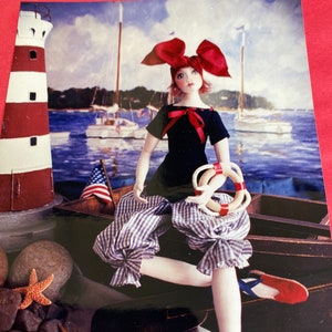 May include: A doll wearing a blue and white gingham dress with a red bow and a red and white life preserver. The doll is sitting in a wooden boat with a red, white, and blue flag in the background.