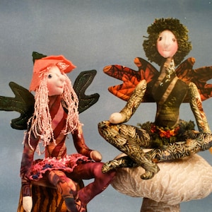 May include: Two handmade fabric dolls with wings, one sitting on a mushroom-shaped cushion and the other on a tree stump. The doll on the mushroom has brown and green patterned clothing and a brown curly wig. The doll on the tree stump has pink and red patterned clothing and a pink wig.