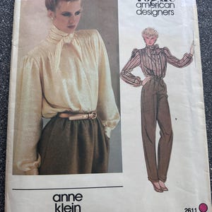 Op de afbeelding: Een Vogue American Designers naaipatroon-envelop voor Anne Klein, nummer 2611. De voorzijde toont een foto van een persoon in een crèmekleurige blouse met lange mouwen en een strik bij de hals, samen met een bruine rok en een mode-illustratie.