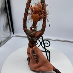 Puede incluir: Una muñeca de tela hecha a mano con un vestido estampado en marrón y verde, una cara marrón y cabello marrón. La muñeca lleva un collar de cuentas y sostiene una cuerda de cuentas en su mano.