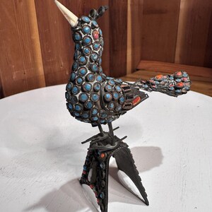 May include: A decorative bird sculpture with a metal frame and a body adorned with blue and orange stones. The bird has a white beak and is perched on a three-legged stand. The sculpture is set against a white surface and a wooden background.