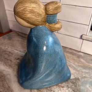 Puede incluir: Una figura de cerámica de una persona con cabello castaño claro peinado en un moño bajo sujeto con una banda azul. La figura viste una prenda azul texturizada y moteada que cae, cubriendo el cuerpo en una postura redondeada y arrodillada.