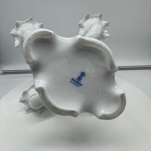 May include: White porcelain candle holder with three candle cups. The holder is decorated with a floral design and has a blue and white mark on the bottom that reads "Kaiser, West Germany".