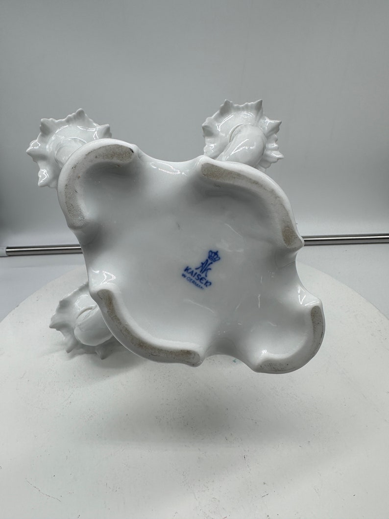 May include: White porcelain candle holder with three candle cups. The holder is decorated with a floral design and has a blue and white mark on the bottom that reads "Kaiser, West Germany".